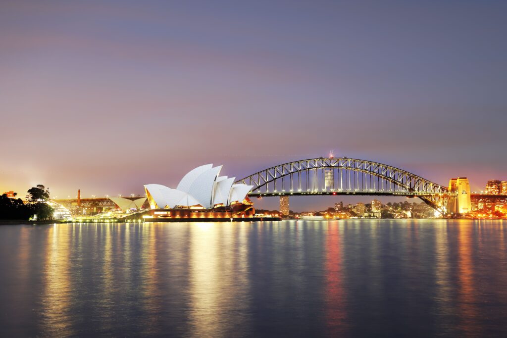 Widok na zatokę w Sydney. Widoczna jest opera oraz Sydney Harbour Bridge. Jest po zachodzie słońca, a od wody odbijają się światła,